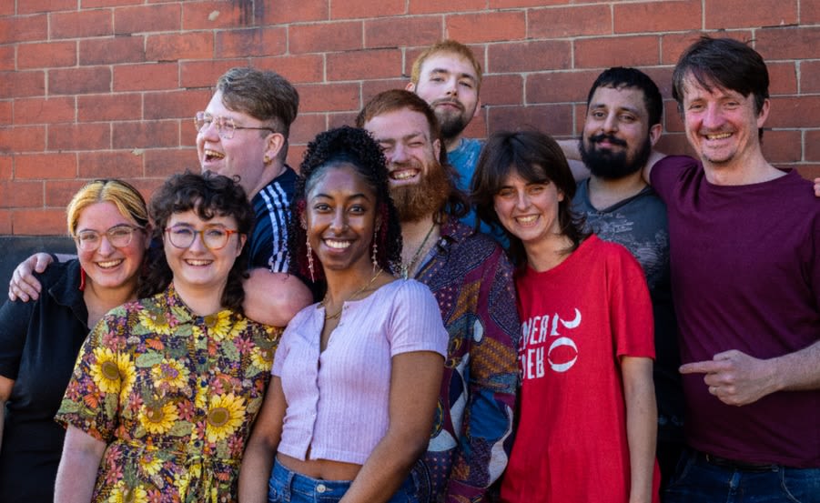 Newcastle Young Producers - a group of eight smiling young people in front of a brick wall with their group facilitator